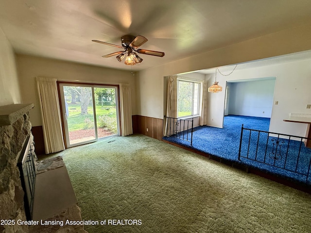 carpeted empty room featuring ceiling fan, healthy amount of natural light, wainscoting, and a stone fireplace