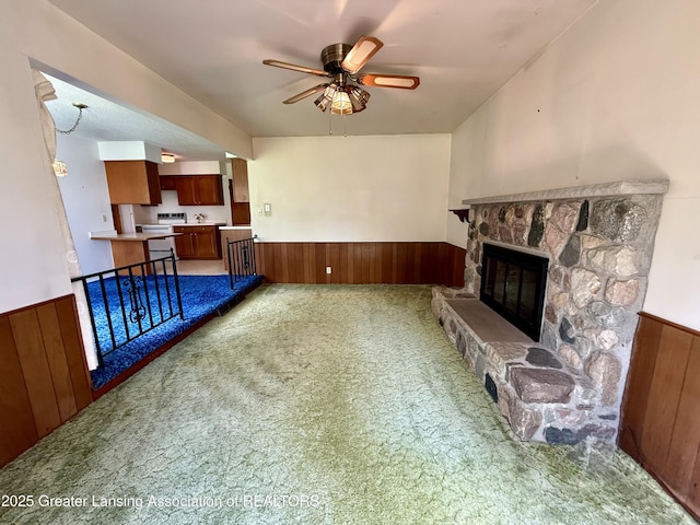 unfurnished living room featuring carpet flooring, wainscoting, a ceiling fan, a stone fireplace, and wood walls