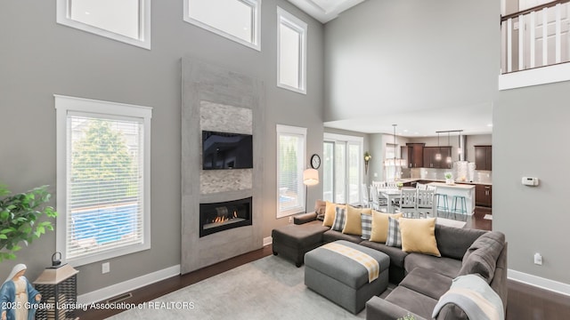 living room with baseboards, dark wood-style flooring, a towering ceiling, a glass covered fireplace, and plenty of natural light