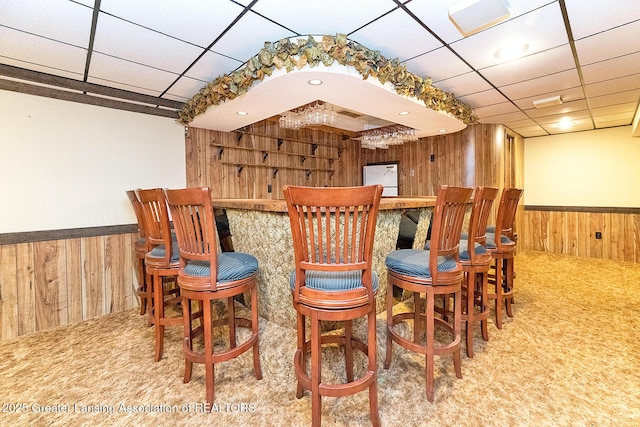 indoor dry bar with wooden walls, wainscoting, carpet, and a drop ceiling