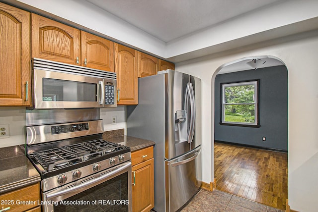 710 Gainsborough Dr, East Lansing, MI 48823 - kitchen with appliances with stainless steel finishes, brown cabinetry, arched walkways, dark stone countertops, and dark wood-style floors