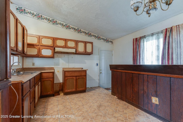 4316 Dumfries Cir, Lansing, MI 48911 - kitchen with a sink, under cabinet range hood, brown cabinets, a chandelier, and glass insert cabinets