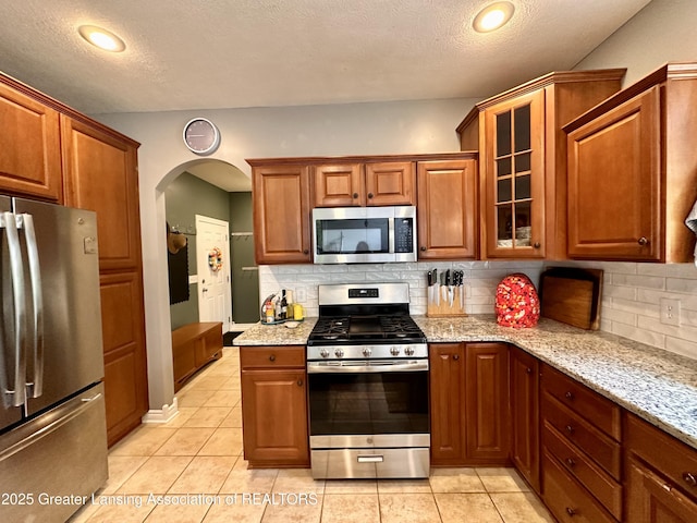 11195 W Herbison Rd, Eagle, MI 48822 - kitchen featuring appliances with stainless steel finishes, arched walkways, light stone counters, glass insert cabinets, and backsplash