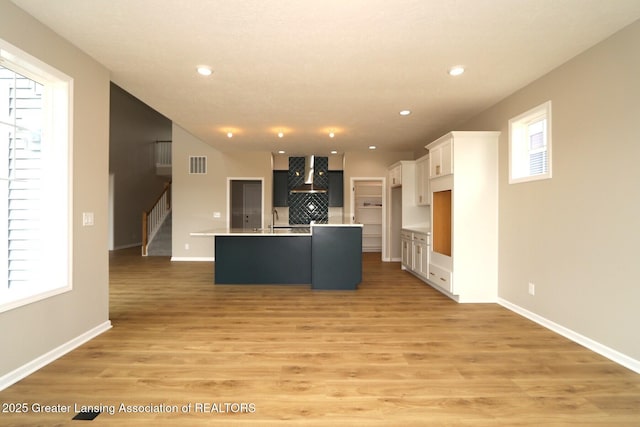 615 Magenta Ct, Haslett, MI 48840 - kitchen with wall chimney exhaust hood, a kitchen island with sink, white cabinets, a sink, and light wood-type flooring