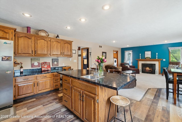 2020 Sunset Dr, Owosso, MI 48867 - kitchen featuring freestanding refrigerator, a kitchen island with sink, a breakfast bar area, brown cabinetry, and a fireplace with flush hearth