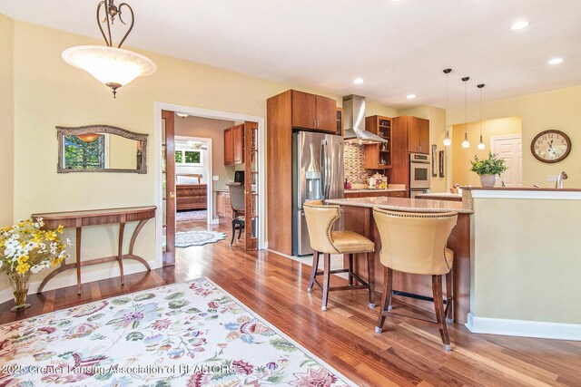 10696 W Clark Rd, Eagle, MI 48822 - kitchen with stainless steel appliances, wall chimney exhaust hood, brown cabinets, wood finished floors, and a breakfast bar