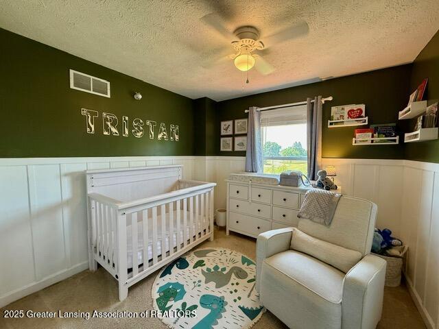 310 Brittany Ln, Mulliken, MI 48861 - carpeted bedroom with a textured ceiling, wainscoting, a crib, a decorative wall, and a ceiling fan