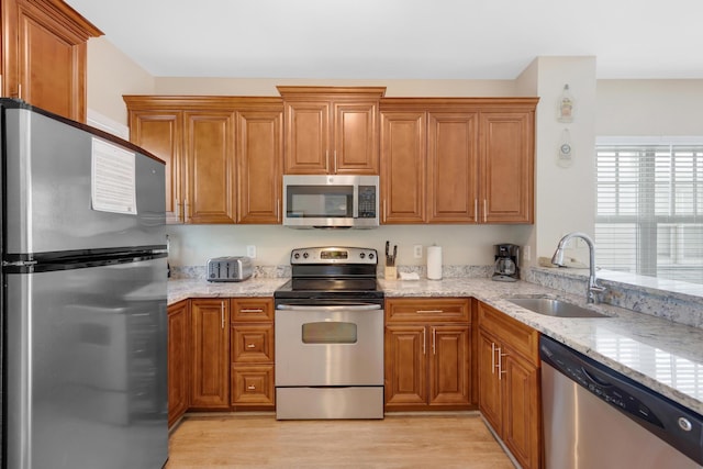 kitchen with light stone countertops, light wood finished floors, a sink, appliances with stainless steel finishes, and brown cabinets