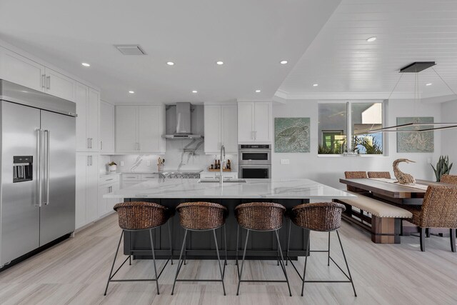 kitchen featuring wall chimney exhaust hood, appliances with stainless steel finishes, light stone counters, white cabinetry, and a sink