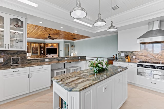 kitchen with a center island, a tray ceiling, glass insert cabinets, wall chimney range hood, and dark stone countertops