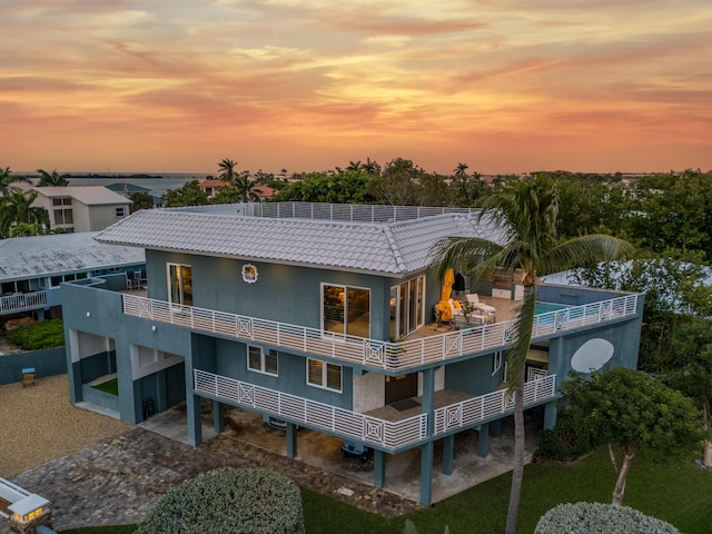 back of property at dusk featuring stucco siding, a balcony, a carport, driveway, and a tiled roof