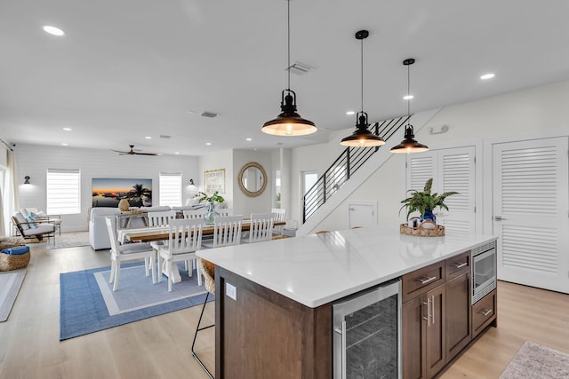 kitchen with hanging light fixtures, ceiling fan, light wood-style floors, wine cooler, and stainless steel microwave