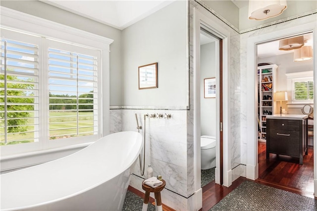 bathroom with a freestanding tub, tile walls, toilet, a wainscoted wall, and wood finished floors