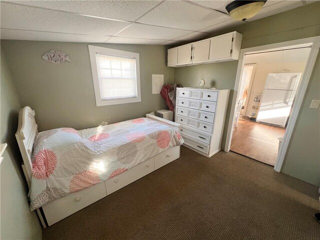 bedroom with a paneled ceiling, dark colored carpet, and white fridge