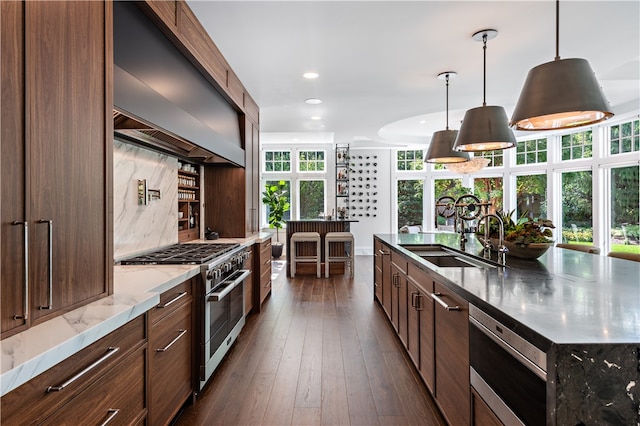 kitchen featuring high end stainless steel range, a kitchen island with sink, decorative light fixtures, sink, and dark wood-type flooring