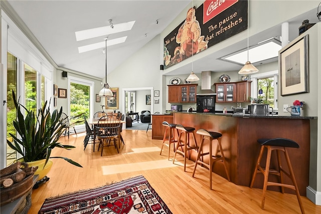 interior space featuring high vaulted ceiling, wall chimney exhaust hood, hanging light fixtures, light hardwood / wood-style flooring, and a skylight