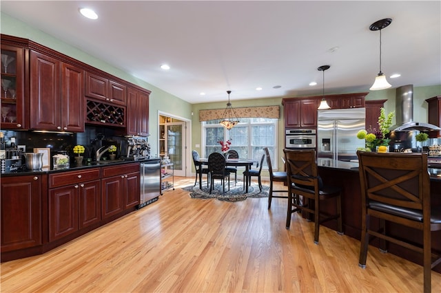 kitchen with decorative light fixtures, wall chimney range hood, a kitchen breakfast bar, stainless steel appliances, and light hardwood / wood-style floors