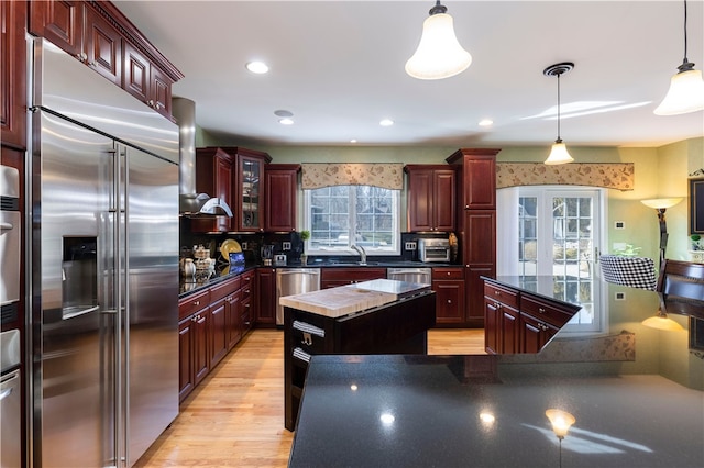 kitchen with appliances with stainless steel finishes, a center island, light hardwood / wood-style flooring, decorative light fixtures, and wall chimney range hood