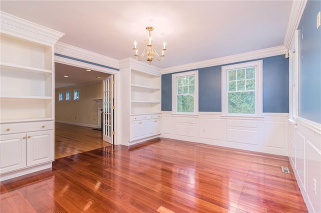 unfurnished dining area featuring a notable chandelier, wainscoting, built in shelves, wood finished floors, and ornamental molding