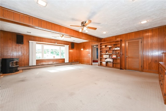 unfurnished living room with ceiling fan, wooden walls, a wood stove, baseboard heating, and light colored carpet