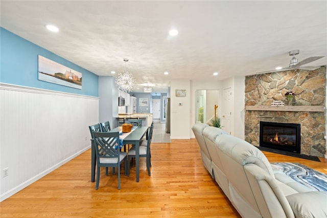 dining area with ceiling fan with notable chandelier, light wood-type flooring, and a stone fireplace