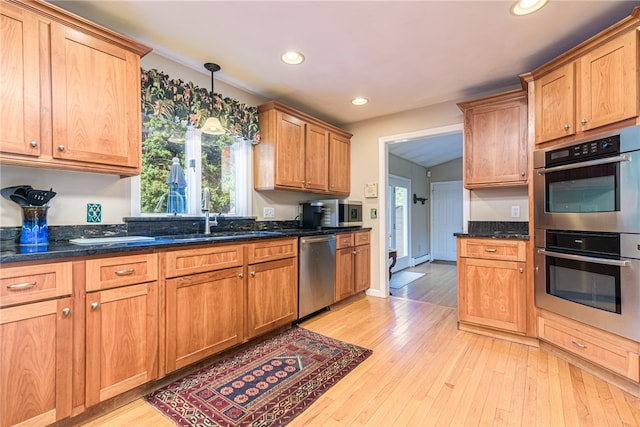 kitchen with appliances with stainless steel finishes, dark stone counters, sink, hanging light fixtures, and light hardwood / wood-style flooring