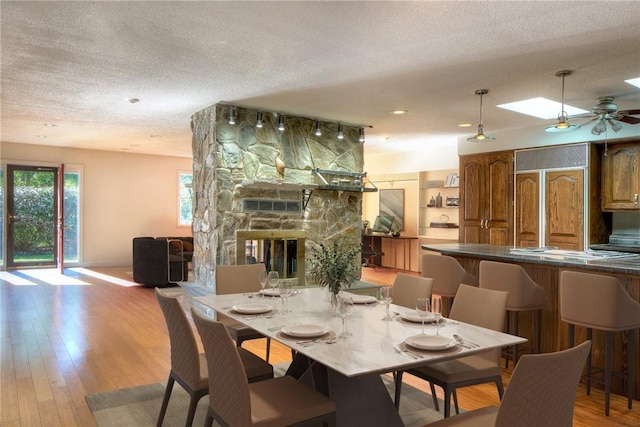 dining area with a textured ceiling, light wood-type flooring, ceiling fan, a skylight, and a stone fireplace