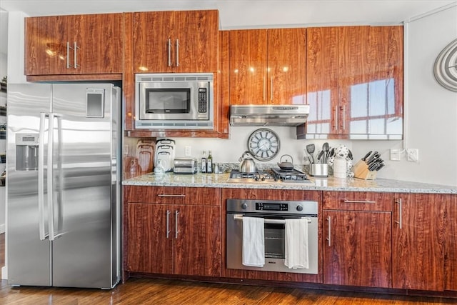 kitchen featuring brown cabinetry, under cabinet range hood, light stone counters, dark wood finished floors, and appliances with stainless steel finishes