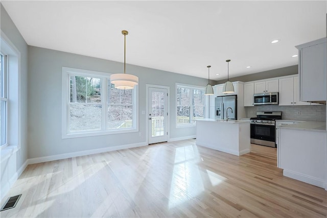 kitchen featuring appliances with stainless steel finishes, decorative light fixtures, tasteful backsplash, an island with sink, and light wood-type flooring