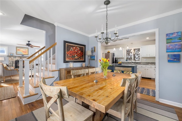 dining area featuring light hardwood / wood-style floors, sink, ornamental molding, ceiling fan with notable chandelier, and a stone fireplace