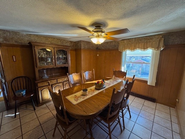 dining space with a textured ceiling, ceiling fan, a baseboard heating unit, wooden walls, and light tile patterned flooring