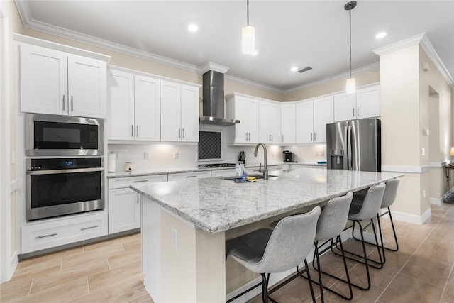 kitchen with stainless steel appliances, a kitchen island with sink, sink, wall chimney range hood, and white cabinetry