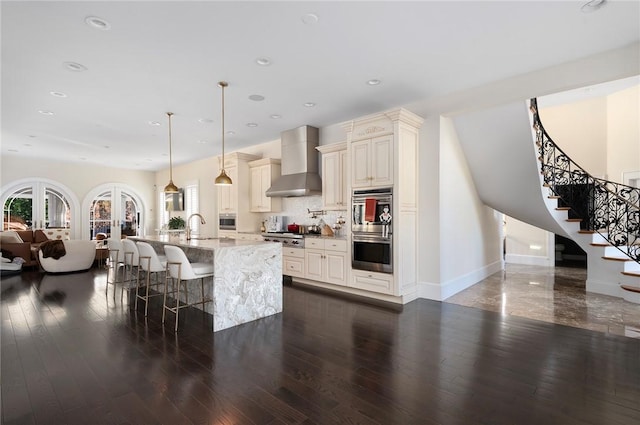 kitchen featuring pendant lighting, wall chimney exhaust hood, decorative backsplash, light stone countertops, and an island with sink