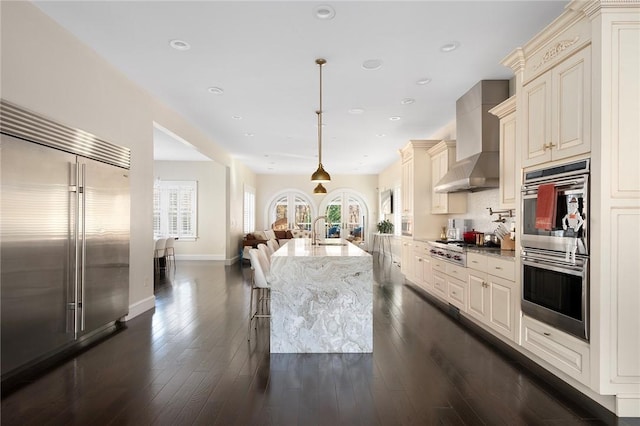 kitchen with appliances with stainless steel finishes, light stone counters, wall chimney exhaust hood, pendant lighting, and cream cabinets