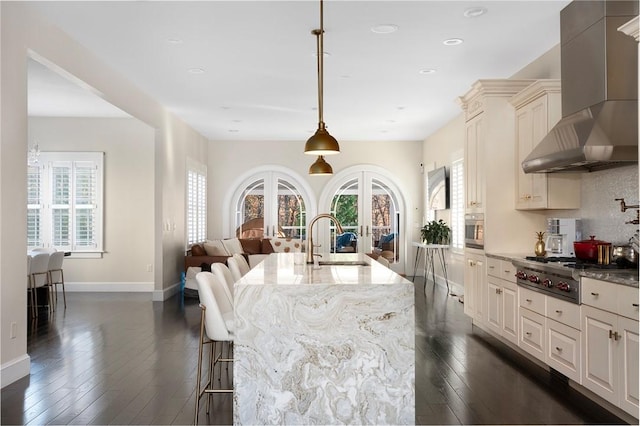 kitchen with french doors, wall chimney exhaust hood, hanging light fixtures, dark stone countertops, and a kitchen island with sink