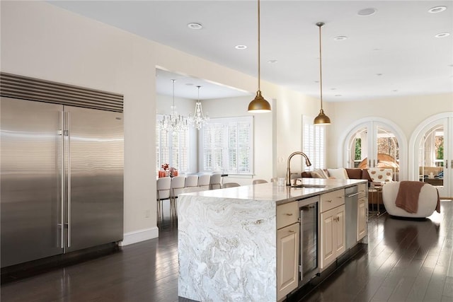 kitchen with pendant lighting, a kitchen island with sink, sink, light stone counters, and stainless steel built in refrigerator