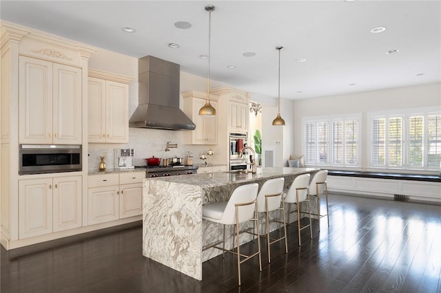 kitchen with dark stone counters, cream cabinets, a center island with sink, wall chimney range hood, and decorative light fixtures