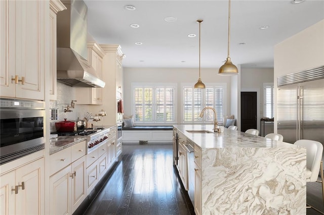 kitchen featuring stainless steel appliances, a breakfast bar area, wall chimney exhaust hood, and an island with sink