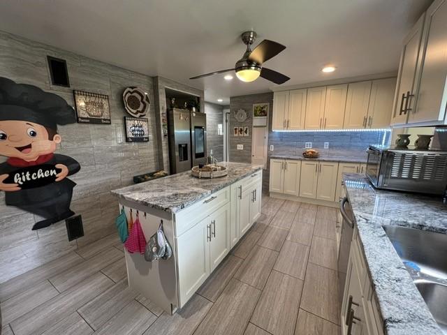 kitchen with ceiling fan, light stone countertops, a kitchen island, stainless steel refrigerator with ice dispenser, and white cabinets