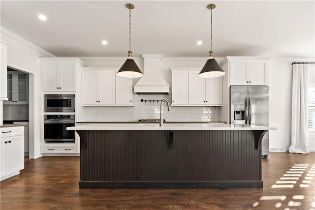 kitchen with appliances with stainless steel finishes, premium range hood, a kitchen island with sink, and pendant lighting