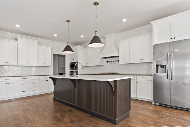 kitchen featuring hanging light fixtures, stainless steel appliances, a center island with sink, white cabinets, and custom range hood