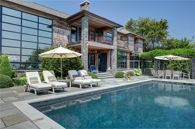 back of property featuring a patio, a balcony, stone siding, an outdoor pool, and a chimney