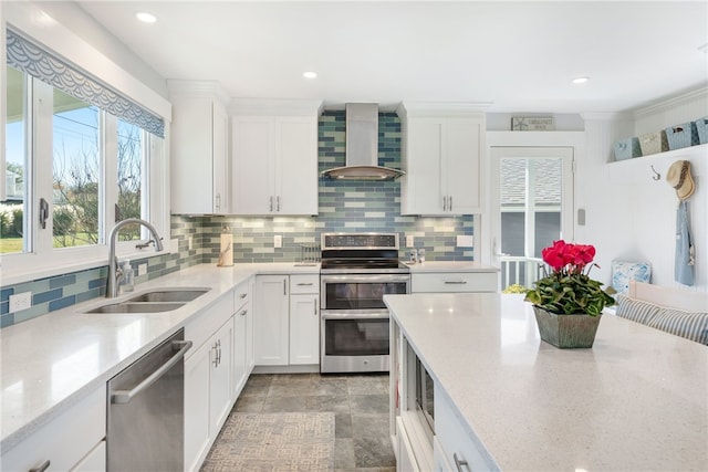 kitchen with light stone countertops, stainless steel appliances, sink, wall chimney range hood, and white cabinetry