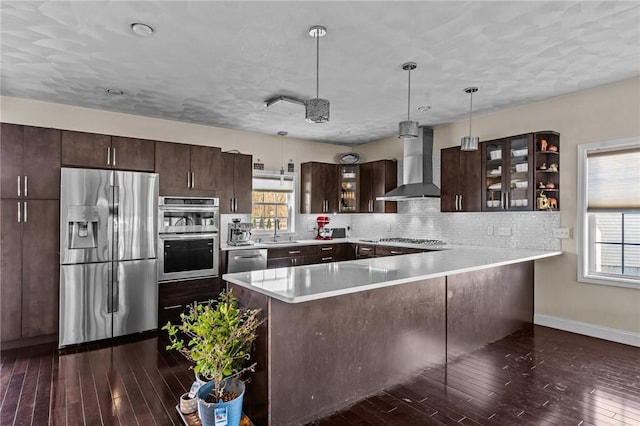 kitchen featuring kitchen peninsula, stainless steel appliances, sink, wall chimney range hood, and hanging light fixtures