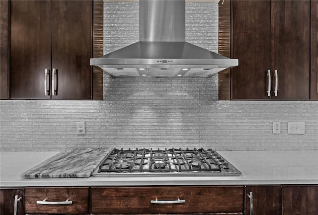 kitchen with decorative backsplash, light stone counters, wall chimney exhaust hood, dark brown cabinetry, and stainless steel gas cooktop