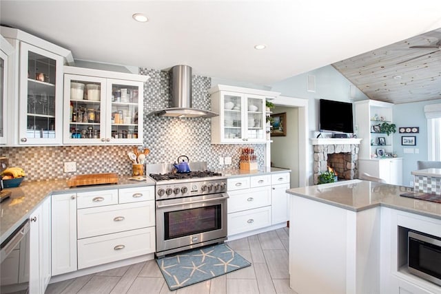 kitchen featuring a fireplace, decorative backsplash, appliances with stainless steel finishes, white cabinets, and wall chimney range hood