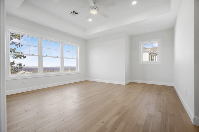 empty room with ceiling fan, light wood-type flooring, a wealth of natural light, and a tray ceiling