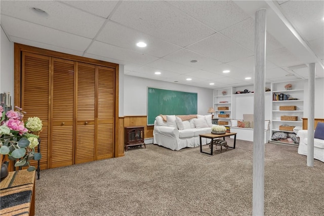 carpeted living room featuring built in shelves, a drop ceiling, wooden walls, and a baseboard radiator