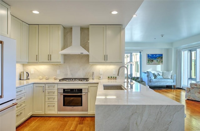 kitchen with wall chimney range hood, tasteful backsplash, white cabinets, appliances with stainless steel finishes, and sink