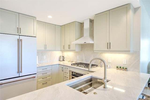 kitchen with light stone countertops, stainless steel appliances, wall chimney range hood, backsplash, and sink